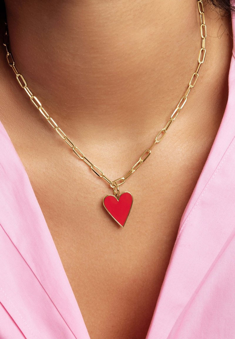 Collar de cadena de oro con un colgante en forma de corazón rojo. La cadena tiene eslabones alargados; el colgante es suave con un acabado brillante. Lleva puesto sobre una tela rosa.