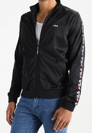 Mand iført sort FILA lynlåstrøje med logo på bryst og ærmer, kombineret med blå jeans, mod en hvid baggrund.