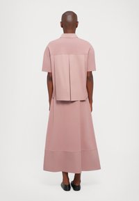 Rosa, kurzärmeliges Hemd mit Rückenschlitz und passendem langen Rock. Glatter Stoff, lockere Passform und subtile Designelemente. Schwarze Ballerinas.