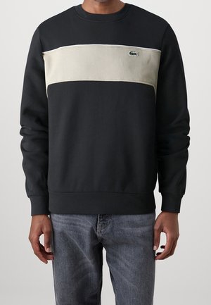 Homme portant un sweat-shirt noir avec un panneau horizontal beige clair sur la poitrine et un petit logo crocodile vert, associé à un jean bleu.