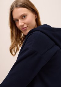 Mörkblå huvjacka med en mjuk textur. Designen har en avslappnad passform och synlig sömnad på axlarna.