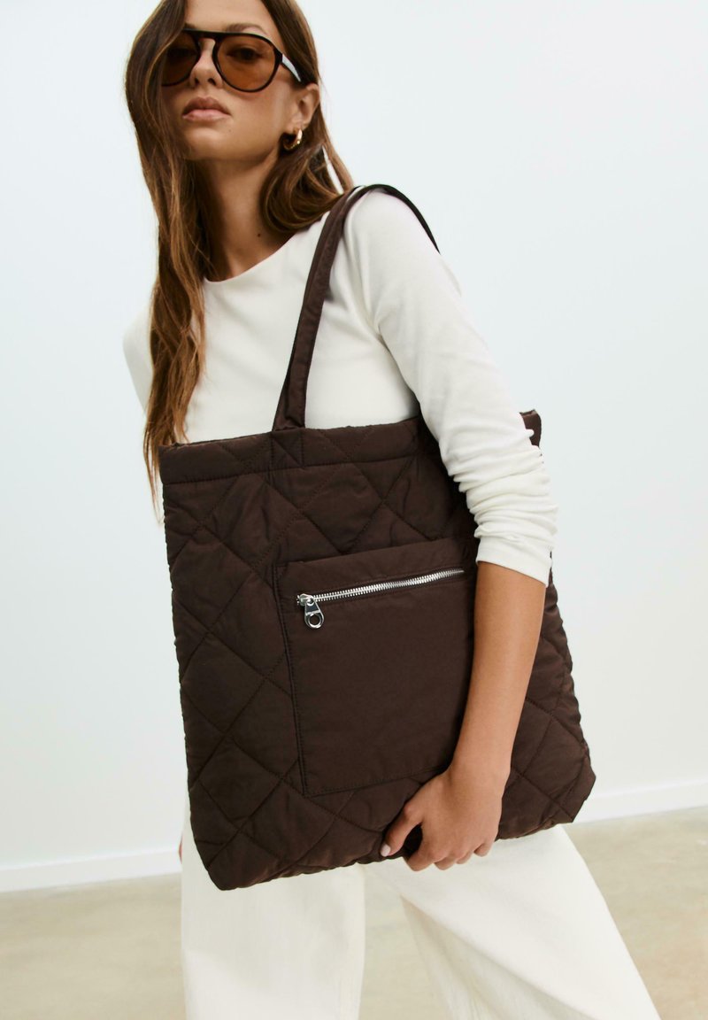 Borsa a tracolla trapuntata marrone con due manici lunghi, dotata di una tasca frontale con cerniera. Texture liscia con un motivo a diamante cucito in tutto il tessuto.
