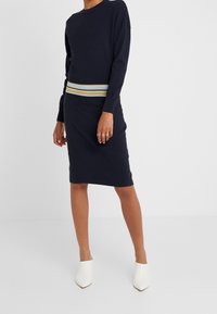 Abito lavorato a maglia di colore blu navy con design a coste, caratterizzato da una cintura multi-righe, maniche lunghe e silhouette aderente. Abbinato a mules bianchi.