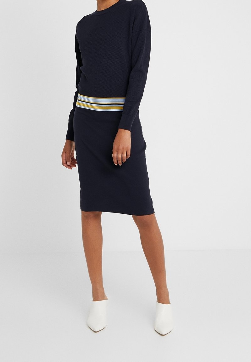Námořnicky modré pletené šaty s žebrovaným vzorem, s vícebarevným proužkovaným páskem, dlouhými rukávy a přiléhavým střihem. Stylizované s bílými mulemi.