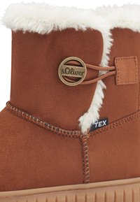 Brązowy zamszowy but z białym sztucznym futrem na brzegu, okrągłym akcentem logo w brązie i kontrastowym przeszyciem wzdłuż szwów. Teksturowana gumowa podeszwa.