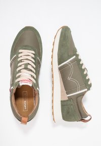 Baskets vertes en daim et tissu, avec des coutures blanches, des lacets beige et une semelle en caoutchouc texturée. Étiquette de logo remarquable sur la languette.