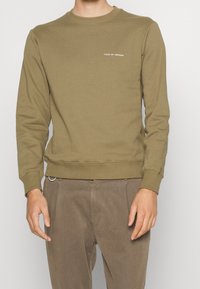 Felpa crewneck verde oliva in un tessuto morbido con polsini e orlo a costine, caratterizzata da un piccolo logo bianco sul petto.
