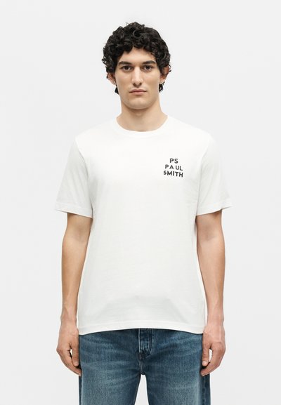 Νέος άνδρας με σγουρά μαλλιά που φοράει λευκό t-shirt PS Paul Smith και μπλε τζιν, στέκεται μπροστά σε ένα απλό φωτεινό φόντο.