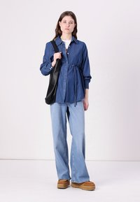 Camisa de denim azul escuro, cingida à cintura, emparelhada com jeans de perna larga azul clara. Bolsa preta de ombro e sapatos de plataforma castanhos.