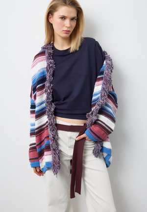 Giovane donna con indosso una maglietta blu navy, pantaloni bianchi con una cintura marrone rossastro e un cardigan multicolore a righe con frange, in piedi contro un muro bianco.