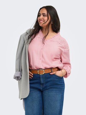 Smilende kvinde i lyserød bluse og blå jeans holder grå blazer over skulderen, med brun bælte og armbånd, på ensartet lys baggrund.
