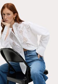 Weißes Hemd mit Knopfleiste und detailliertem Cut-out-Stickerei auf der Vorderseite, kombiniert mit blauen Jeans. Modell sitzt auf einem schwarzen Stuhl.