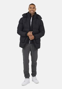 Man die glimlacht en een zwarte pufferjack, grijze broek en witte sneakers draagt terwijl hij tegen een witte achtergrond staat.