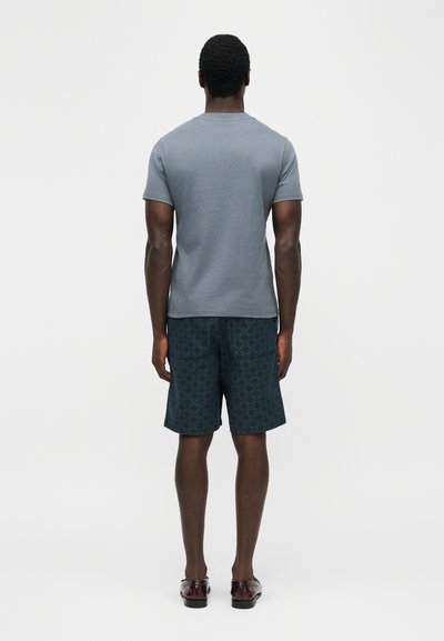 Άνδρας που στέκεται με την πλάτη προς τα εμπρός, φοράει γκρι κοντομάνικο πουκάμισο, σκούρα σορτς με σχέδια και πίσω τσέπες, και καφέ παπούτσια.