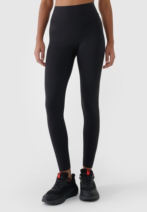 Osoba ubrana w czarne legginsy z wysokim stanem oraz czarne buty sportowe z czerwonymi pętlami, stojąca na jednolitym jasnym tle.