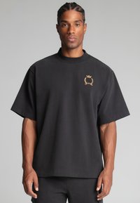 Sort oversized t-shirt lavet af bomuld, med korte ærmer, høj hals og et guld broderet logo på venstre bryst.