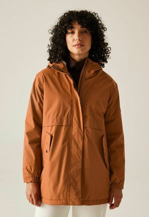 Kobieta o kręconych włosach, ubrana w rdzawą, wodoodporną kurtkę z kapturem, posiadającą kieszenie z klapami i elastyczne mankiety, stojąca na jednolitym jasnym tle.