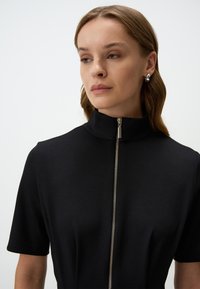 Abito nero a maniche corte con colletto alto e dettaglio con cerniera argentata, con una texture liscia e una vestibilità sartoriale.