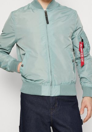 Person trägt eine hellgrüne Bomberjacke mit gerippten Bündchen und Kragen, Reißverschluss vorne, Seitentaschen und rotem "Remove Before Flight"-Anhänger an der Ärmeltasche.