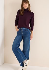 Maglione a collo alto bordeaux, jeans a gamba larga blu e sneakers bianche con dettagli neri. Tessuto a maglia e vestibilità casual. Sfondo semplice.