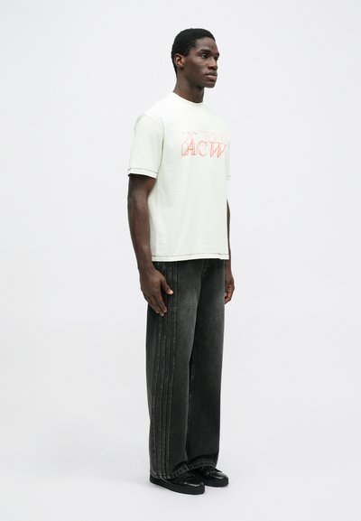 Camiseta de algodón verde claro con texto gráfico en rojo, combinada con jeans de mezclilla de pierna ancha gris oscuro con costuras verticales y zapatillas negras.