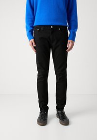 Jeans negros ajustados con cinco bolsillos, herrajes metálicos y costuras blancas, combinados con un suéter de punto azul y zapatillas negras.