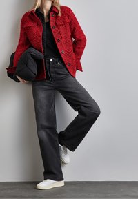 Red textured jacket with black buttons, paired with high-waisted black jeans. White sneakers complete the outfit. Grey background.