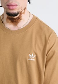 Tricou din bumbac maro cu decolteu rotund și mâneci scurte. Prezentă un logo Adidas brodat alb în zona stângă a pieptului.