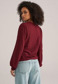 Žena s kovrčavom kosom koja nosi teksturiranu bordo bluzu dugih rukava i svijetloplave traperice, okreće se preko ramena.