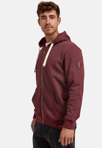 Bordeauxroter Hoodie aus strukturiertem Stoff, mit einem Frontreißverschluss, Kordelzugkapuze, seitlichen Taschen und einem Logo-Patch am Ärmel.