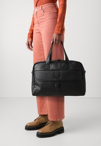 Bolso tote de cuero negro con dos asas, cierre de cremallera y bolsillo delantero con tapa. Textura suave con detalles de costura en contraste.