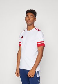 Weißes Sportshirt mit roten Streifen an den Ärmeln und auf der Schulter, aus glattem Stoff. Verfügt über drei rote Streifen auf beiden Schultern.