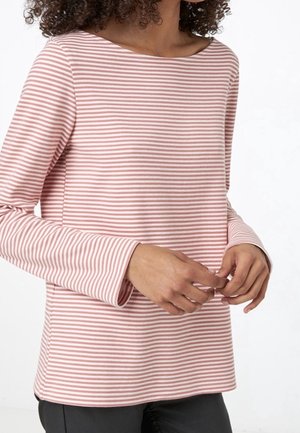 Femme portant un haut à manches longues à rayures horizontales roses et blanches, les mains jointes légèrement devant elle, avec des cheveux bouclés partiellement visibles.