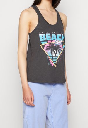 Vrouw draagt een donkergrijze tanktop met een kleurrijke "Palm Springs Beach Club" afbeelding en lichtblauwe broek, staand tegen een effen achtergrond.