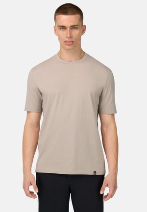 T-shirt beige à manches courtes en tissu doux, avec un col rond et une petite étiquette sur le côté inférieur, porté avec un pantalon foncé.