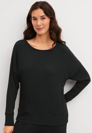Femme aux longs cheveux bruns portant un haut noir côtelé à manches longues et ample, debout devant un fond blanc uni.