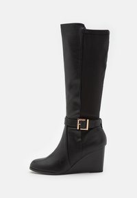 Botte à talon compensé noire mi-mollet avec une boucle carrée dorée sur la sangle latérale, devant en cuir lisse et panneau arrière en tissu extensible.