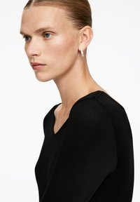 Junge Frau mit blauen Augen und zurückgegeltem Haar, die ein schwarzes Oberteil und kleine silberne Creolen trägt, und nach links gegen einen weißen Hintergrund schaut.