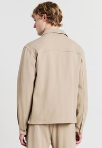 Giacca beige, strutturata, con colletto e maniche lunghe. Presenta una linea dritta e dettagli di cucitura sottili. Polsini con un bottone.