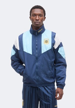Homme portant une veste de sport Adidas bleue et bleu clair et un pantalon assorti avec le logo AFA, debout avec une main dans la poche devant un fond uni.
