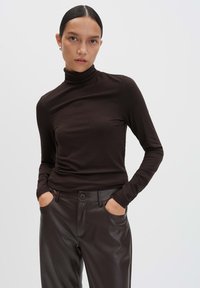Bruine turtleneck top met lange mouwen van een zachte, rekbare stof. Het model draagt het met high-waisted, donkerbruine leren broek.