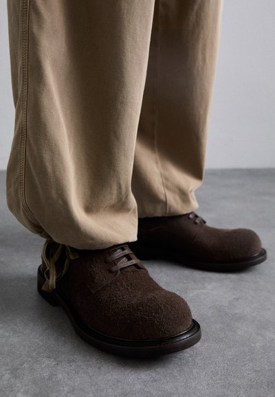Zapatos de fieltro marrón con puntera redondeada, cordones planos y una suela robusta, combinados con pantalones beiges, situados sobre una superficie gris.