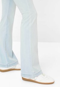 Persona con jeans a zampa di colore azzurro chiaro e sneakers bianche con suola in gomma, in piedi davanti a uno sfondo bianco uniforme.