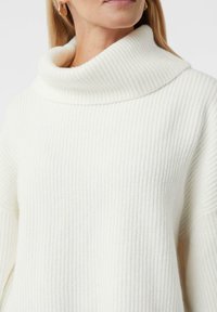 Kobieta w białym prążkowanym swetrze z golfem i złotymi kolczykami w kształcie kół, odsłaniająca szyję i górną część klatki piersiowej.