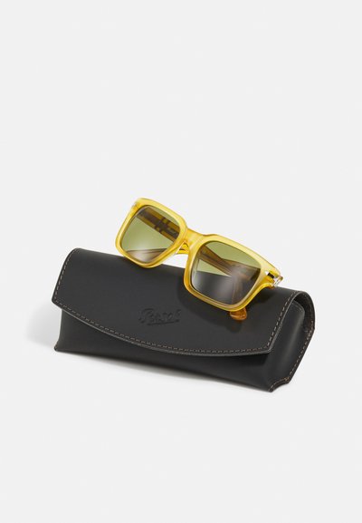 Ochelari de soare dreptunghiulari galbeni cu lentile verzi, așezați pe un toc din piele neagră cu cusături maro și logo embosat.