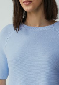 Pull en maille bleu clair à manches courtes et col rond. Le tissu texturé présente un détail en côte subtil aux poignets et à l'ourlet.