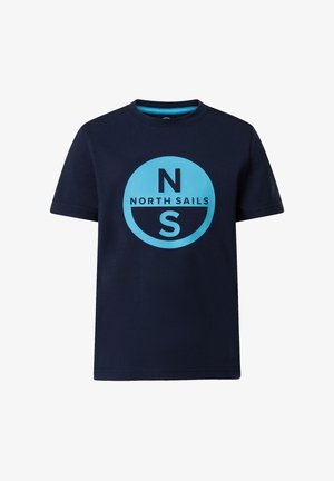 Jeune garçon debout, les mains dans les poches, portant un t-shirt North Sails bleu marine, un pantalon blanc et des baskets blanches sur fond uni.