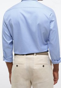 Světle modrá a bílá košile se vzorem káro, s vyhrnutými rukávy, doplněná světle béžovými kalhotami s kapsami vzadu a knoflíkovými detaily.