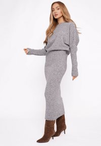 Szary dzianinowy komplet dwuczęściowy z luźnym swetrem z długimi prążkowanymi rękawami oraz dopasowaną spódnicą kończącą się poniżej kolan, w połączeniu z brązowymi kozakami.