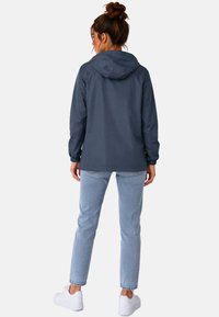 Vrouw met bruin haar in een knot, gekleed in een donkerblauwe capuchonjas, lichtblauwe spijkerbroek en witte sneakers, staande met de rug naar de camera op een witte achtergrond.